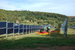 das Photo zeigt einen Traktor, der zwischen zwei Reihen von Photovoltaikmodulen eine Wiese mäht.